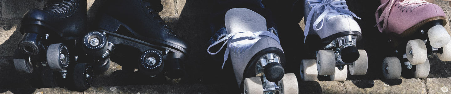 Three pairs of Rookie roller skates lined up on concrete, featuring different colors—black, white, and pink. They are lit by sunlight.